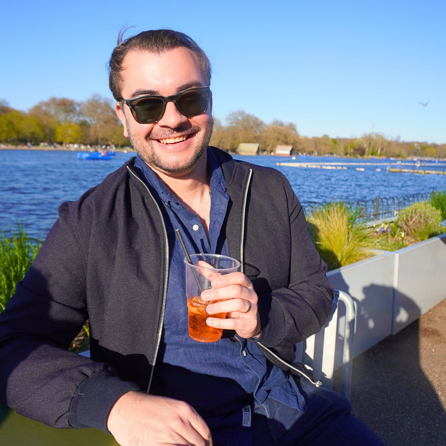 Man sitting by a lake holding a drink, wearing sunglasses and a dark jacket.