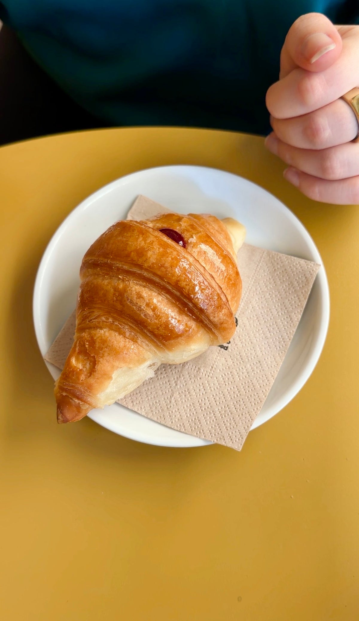 A golden-brown croissant on a white plate with a beige napkin, set on a yellow table with a person's hand visible in the upper right corner.
