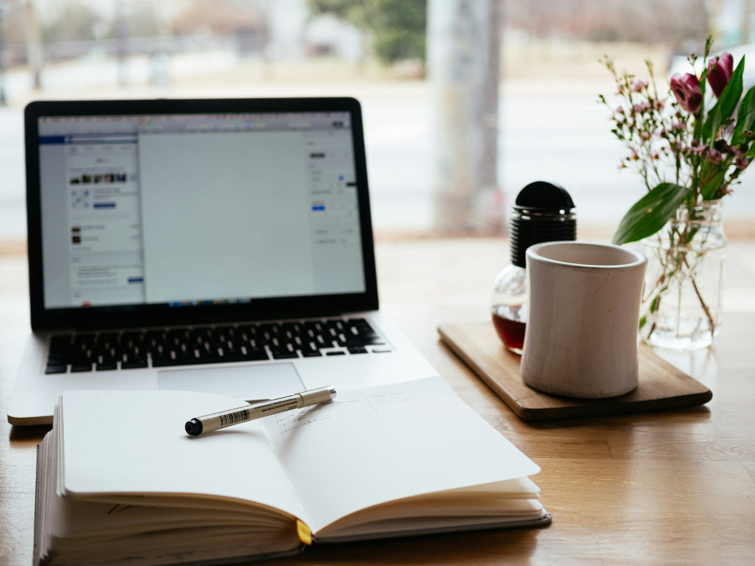 Laptop, notebook, pen, and mug on a wooden desk with a blurred background
