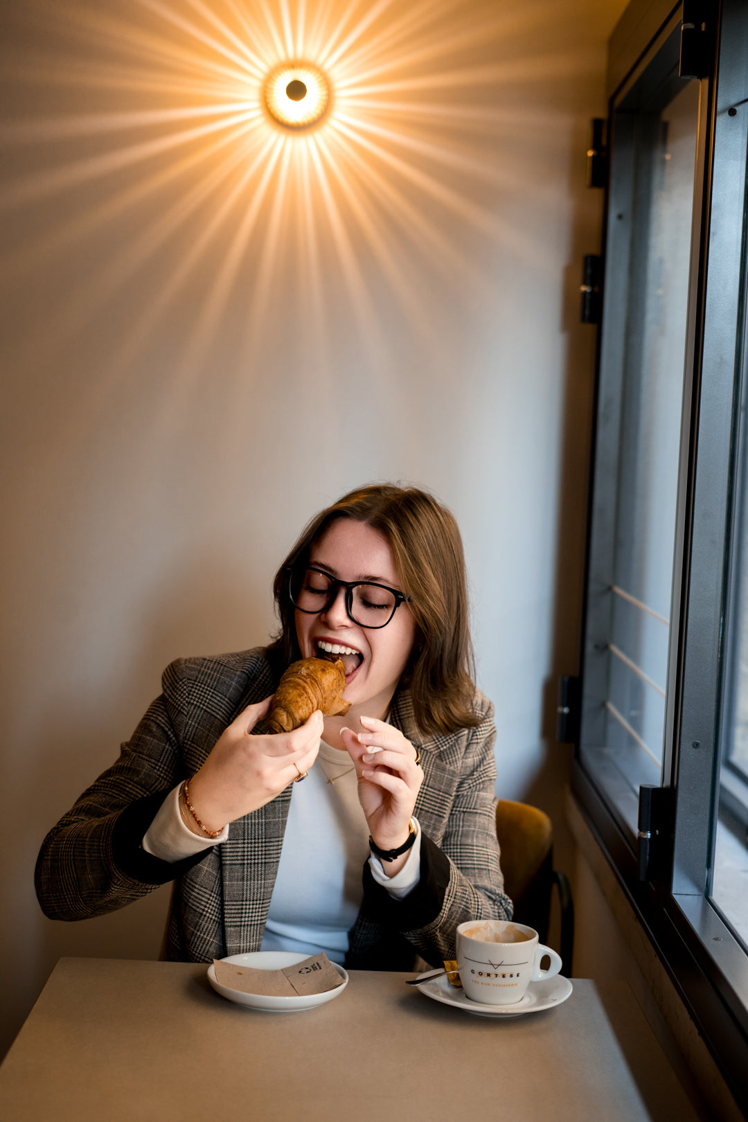 A woman with glasses is sitting at a cafe table, eating a croissant and smiling. A cup of coffee sits on the table in front of her, with a warm sunburst light glowing on the wall behind her.